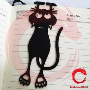 Pode incluir: Marcador de livros preto em forma de gato, feito de material cortado a laser. O marcador apresenta uma silhueta de gato com um rosto sorridente, bigodes e patas. O marcador é inserido em um caderno com uma seção de memorando.