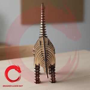 May include: A laser-cut wooden dinosaur model, assembled from multiple layers. The dinosaur is light brown with dark brown detailing, with a long neck and tail. The model stands on four legs. The text "DRAGON LASER CUT" is visible.