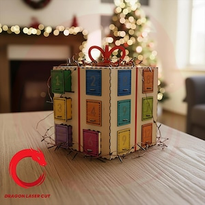 Könnte beinhalten: Ein quadratischer Adventskalender aus Holz in Form eines Geschenkkartons mit einer roten Schleife oben. Die Box hat zwölf kleine, bunte Türen, die von eins bis zwölf nummeriert sind und jeweils einen Metallgriff haben. Dünne Lichterketten umwickeln die Box.