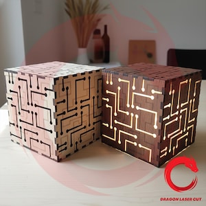 Puede incluir: Dos lámparas de madera con forma de cubo con diseños de circuitos. Una lámpara es de color claro con líneas negras, la otra es marrón oscuro con líneas iluminadas. Las lámparas están sobre una superficie clara.