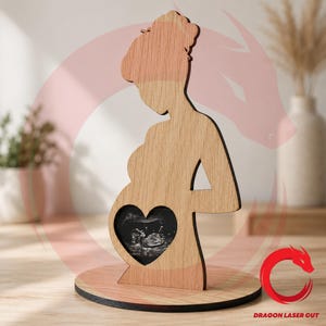 Può includere: Silhouette in legno di una persona incinta con un ritaglio a forma di cuore che mostra un'immagine ecografica. La figura è realizzata in legno chiaro e poggia su una base rotonda. Il design è un oggetto decorativo, forse un ricordo o un regalo.