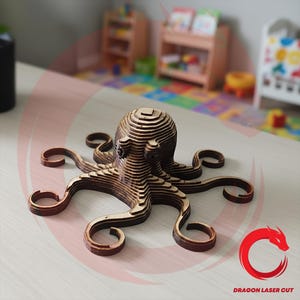 Könnte beinhalten: Eine Holzskulptur eines Oktopus mit einem geschichteten Design. Der Oktopus hat acht gekräuselte Tentakel und einen abgerundeten Kopf. Die Skulptur besteht aus hellem und dunklem braunem Holz und steht auf einer hellen Oberfläche. Das Logo "DRAGON LASER CUT" befindet sich unten rechts.