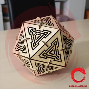 May include: A wooden geometric puzzle in the shape of a dodecahedron. The puzzle has intricate laser-cut designs with black accents on each face. The interlocking pieces are held together with small wooden pegs. The overall color is a light tan.