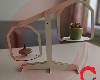 May include: A light-colored wooden balance scale featuring two hanging platforms. A small brown wooden spinning top sits on the left platform, while a green succulent in a two-toned green ceramic pot rests on the right platform. The structure is laser-cut.