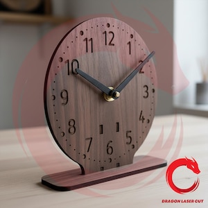May include: A circular dark brown wooden desk clock featuring laser-cut numerals from 1 to 12. The clock has two black hands and a small gold-colored center pin. It is mounted on a rectangular wooden base, providing a stable upright position.