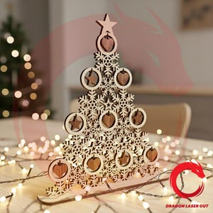 Peut inclure: Décoration de sapin de Noël en bois avec des découpes de flocons de neige et d'étoiles. L'arbre présente des ornements en forme de cœur et d'étoile. L'arbre est en bois clair avec des accents de bois plus foncés. La base est un traîneau en bois.