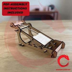 Puede incluir: Maqueta de catapulta de madera de color marrón claro. El modelo tiene una base rectangular, un brazo en ángulo y una pequeña plataforma. El texto "PDF-ASSEMBLY INSTRUCTIONS INCLUDED" está incluido. El logotipo de Dragon Laser Cut está en la esquina inferior derecha.