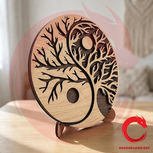 May include: A circular wooden wall art piece featuring a laser-cut yin and yang design formed by two interlocking trees. The light-colored wood contrasts with the dark-colored wood, and the piece is supported by two small wooden stands.