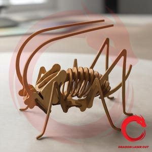 Può includere: Modello di cavalletta in legno, tagliato al laser e assemblato. L'insetto presenta un corpo, zampe e antenne dettagliati. Il modello è realizzato in legno marrone chiaro ed è esposto su una superficie chiara. Il testo "DRAGON LASER CUT" è visibile.