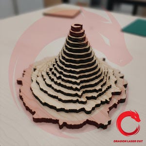 Op de afbeelding: Een gelaagde houten sculptuur van een berg of vulkaan. De structuur is gemaakt van lichtgekleurd hout met donkere, verbrande randen, waardoor een gestreept effect ontstaat. De basis is lichtbruin van kleur en de top is een donkerdere bruintint. Waarschijnlijk lasergesneden.