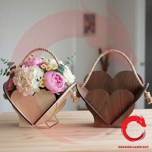 May include: Two wooden heart-shaped baskets with rope handles. One basket is filled with pink, white, and yellow flowers. The other basket is empty. The baskets are on a wooden table. The text "DRAGON LASER CUT" is visible.