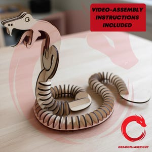 Puede incluir: Un rompecabezas de serpiente articulada de madera clara con la cabeza levantada y el cuerpo enroscado. El diseño presenta segmentos cortados con láser. Un gráfico rectangular rojo muestra el texto VIDEO-ASSEMBLY INSTRUCTIONS INCLUDED.