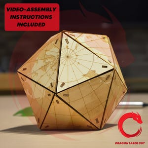 Op de afbeelding: Een geometrische houten puzzel in de vorm van een veelvlak, met een wereldkaartontwerp. De puzzel is gemaakt van lichtgekleurd hout en heeft een complexe, veelzijdige structuur. De tekst op de afbeelding luidt "VIDEO-ASSEMBLY INSTRUCTIONS INCLUDED". De diameter is ongeveer 20 cm.