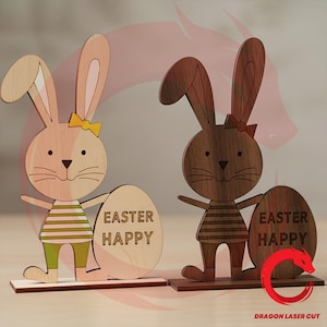 May include: Two wooden Easter bunny ornaments. One is light-coloured with a yellow bow and green trousers, the other is dark brown with a red bow. Each bunny holds an egg with "EASTER HAPPY" engraved. They stand on rectangular bases.