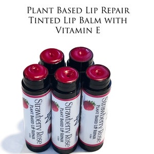 Könnte beinhalten: Sechs Tuben getönten Lippenbalsams mit Erdbeer-Rosen-Duft und pflanzlicher Lippenreparaturformel. Die Lippenbalsam-Tuben sind schwarz mit weißem Text und einer Erdbeergrafik.