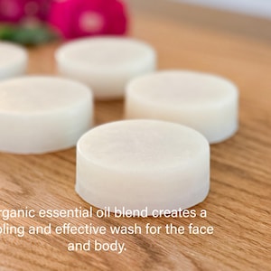 Five organic cream colored face and body wash soaps  layed out on a wood counter in front of red roses and plantain.
