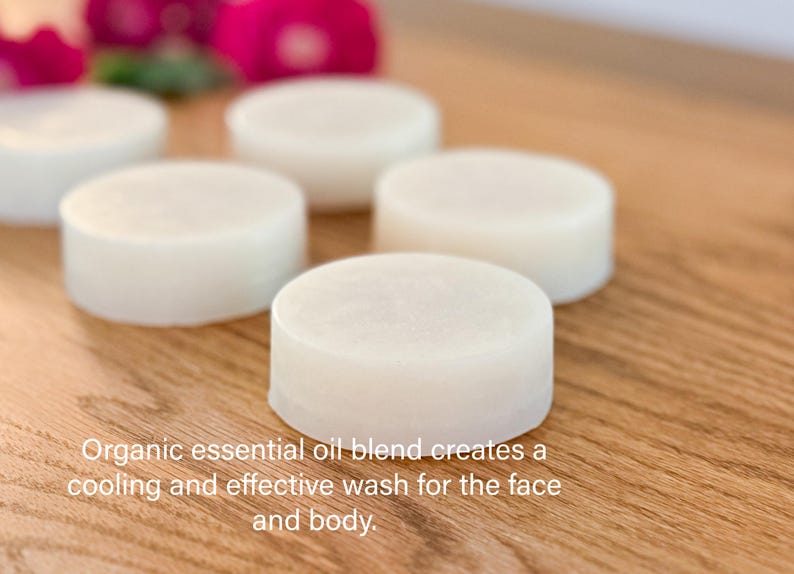 Five organic cream colored face and body wash soaps  layed out on a wood counter in front of red roses and plantain.
