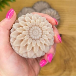 May include: A light beige, flower-shaped soap bar held in a hand with bright pink fingernails. The soap has a detailed floral design with a central spiral. Other similar soaps are visible in the background, suggesting a set or collection.