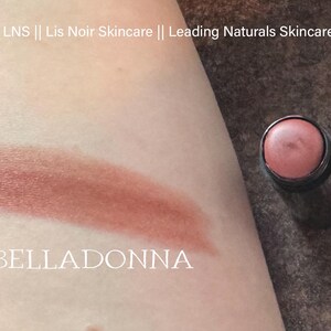 K&ouml;nnte beinhalten: Nahaufnahme eines braunen Creme-Rouge-Sticks mit dem Wort "BELLADONNA" auf der Haut unter dem Farbmuster. Das Creme-Rouge befindet sich in einem schwarzen Beh&auml;lter.