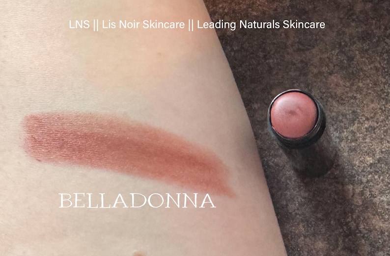 K&ouml;nnte beinhalten: Nahaufnahme eines braunen Creme-Rouge-Sticks mit dem Wort "BELLADONNA" auf der Haut unter dem Farbmuster. Das Creme-Rouge befindet sich in einem schwarzen Beh&auml;lter.