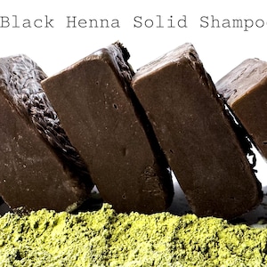 Pode incluir: Close-up de várias barras de champô sólido retangulares castanhas escuras encostadas umas às outras. As barras estão sobre uma cama de pó verde. O texto "Black Henna Solid Shampoo" está no topo.