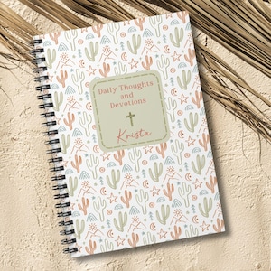May include: Spiral-bound notebook with a white cover featuring a repeating pattern of cacti, stars, and mountains in muted colors. The center has a label that reads "Daily Thoughts and Devotions" with a cross and the name "Krista".
