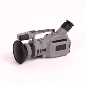 Può includere: Una piccola videocamera grigia e nera con obiettivo, maniglia e mirino. La fotocamera ha un design rettangolare ed è posta su uno sfondo bianco. L'obiettivo è nero e il corpo è grigio.