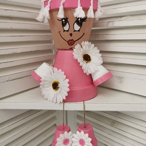 Può includere: Un vaso di terracotta dipinto di rosa con un bordo bianco e frange, dipinto con un viso sorridente e occhi grandi. Il vaso è decorato con fiori bianchi e ha due vasi rosa più piccoli appesi ad esso.