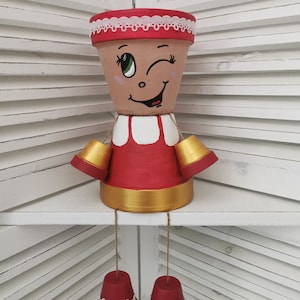 Könnte beinhalten: Ein skurriler Pflanzer in Form einer Person mit einem zwinkernden Gesicht, roten und weißen Kleidungsstücken und goldenen Akzenten. Der Pflanzer hat eine rote Basis mit weißer Spitzenverzierung und goldenen Akzenten.