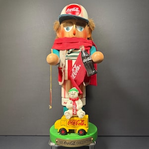 May include: A Coca-Cola themed nutcracker figurine. The figurine wears a Coca-Cola cap and scarf, holding a Coca-Cola bottle and flag. A polar bear sits on a yellow Coca-Cola truck. The base reads "THE Coca-Cola COLLECTOR."