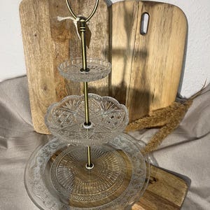 Könnte beinhalten: Ein dreistufiger Servierständer mit klaren Glasplatten und einem goldfarbenen Metallrahmen. Der obere Griff ist herzförmig. Der Ständer wird vor Holzschneidebrettern präsentiert.