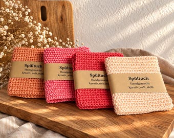 Può includere: Quattro strofinacci quadrati all'uncinetto nei toni pesca, rosa, rosso e crema. Ogni panno è avvolto in una fascia di carta marrone con il testo Spültuch, Handgemacht e kreativ_welt_stefii, disposti in fila su una superficie di legno.