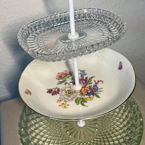 Può includere: Un alzatina a tre livelli con struttura in metallo bianco. Il livello superiore è in vetro trasparente, il livello intermedio è un piatto bianco con un motivo floreale e il livello inferiore è in vetro verde.