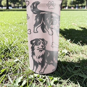K&ouml;nnte beinhalten: Ein rosa Becher mit schwarz-wei&szlig;en Illustrationen von Hunden, Pfotenabdr&uuml;cken und einem Knochen. Der Becher hat ein gl&auml;nzendes Finish und steht auf gr&uuml;nem Gras.
