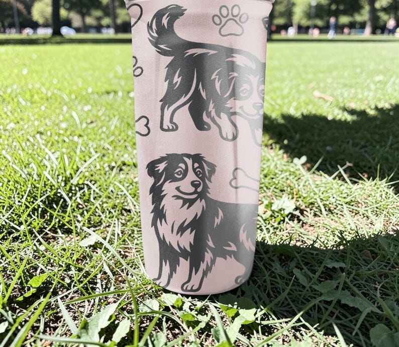 K&ouml;nnte beinhalten: Ein rosa Becher mit schwarz-wei&szlig;en Illustrationen von Hunden, Pfotenabdr&uuml;cken und einem Knochen. Der Becher hat ein gl&auml;nzendes Finish und steht auf gr&uuml;nem Gras.