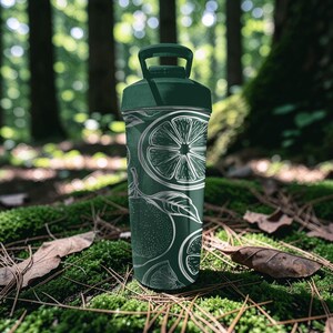 May include: A dark green water bottle with a white citrus fruit design. The bottle has a handle on the lid and is cylindrical in shape. The design includes slices of citrus fruit and leaves. The bottle is set against a natural background of moss and trees.