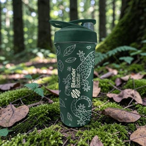 May include: A dark green Blender Bottle with a white lizard and leaf pattern. The bottle has a dark green lid and a clear spout. The Blender Bottle logo is visible on the side. The bottle is designed for mixing protein shakes and other beverages.