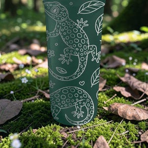 May include: A dark green tumbler featuring a white outline pattern of geckos, leaves, and flowers. The design is repeated around the tumbler. The tumbler is cylindrical and appears to be made of a durable material.