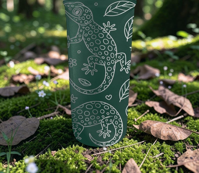 May include: A dark green tumbler featuring a white outline pattern of geckos, leaves, and flowers. The design is repeated around the tumbler. The tumbler is cylindrical and appears to be made of a durable material.