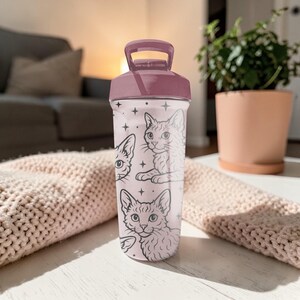 May include: A pink and white water bottle with a dark pink lid and handle. The bottle features a pattern of cat illustrations and stars. The bottle is sitting on a white surface.
