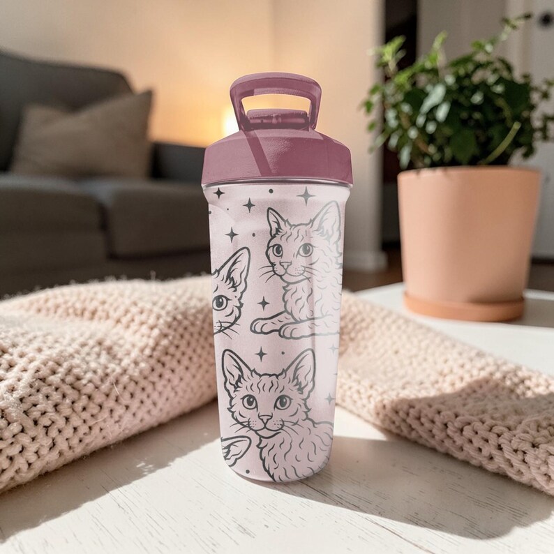 May include: A pink and white water bottle with a dark pink lid and handle. The bottle features a pattern of cat illustrations and stars. The bottle is sitting on a white surface.