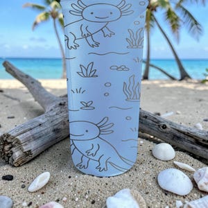 May include: A light blue tumbler featuring a repeating axolotl design with aquatic plants and bubbles. The axolotls are drawn in a simple, cartoon style. The tumbler is on a sandy beach with driftwood and seashells.
