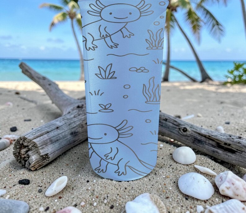 May include: A light blue tumbler featuring a repeating axolotl design with aquatic plants and bubbles. The axolotls are drawn in a simple, cartoon style. The tumbler is on a sandy beach with driftwood and seashells.