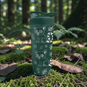 May include: A dark green water bottle with a white lemur and leaf pattern. The bottle has a flip-top lid and is set against a backdrop of green moss and foliage. The lemur design includes a striped tail.