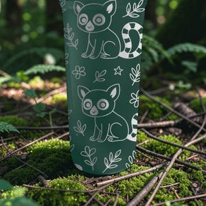 May include: A dark green tumbler featuring a repeating pattern of lemur illustrations and botanical elements in white. The lemurs have large eyes and striped tails. The design includes flowers, stars, and leaves.
