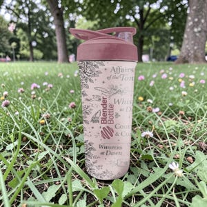 Pode incluir: Uma garrafa Blender rosa com tampa a condizer, contra um fundo de relva verde e flores. A garrafa apresenta desenhos florais e de escrita, com o texto "Affairs of the Ton" e "Whispers at Dawn."