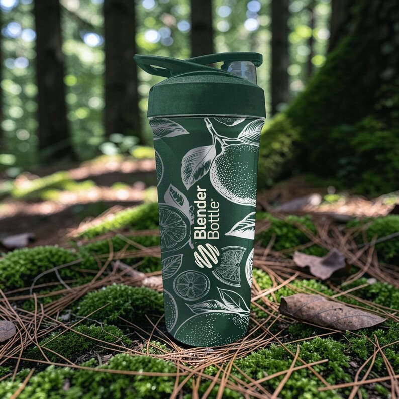 May include: A green Blender Bottle with a white citrus fruit design. The bottle has a dark green lid and a clear body. The text "Blender Bottle" is printed on the side. The bottle is sitting on a bed of moss and pine needles.