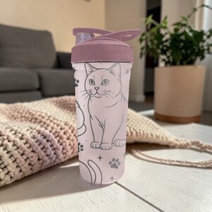 May include: A pink and mauve water bottle featuring a cat illustration. The bottle has a flip-top lid and a light pink body with a cat design, paw prints, and stars. The lid is a darker mauve color.