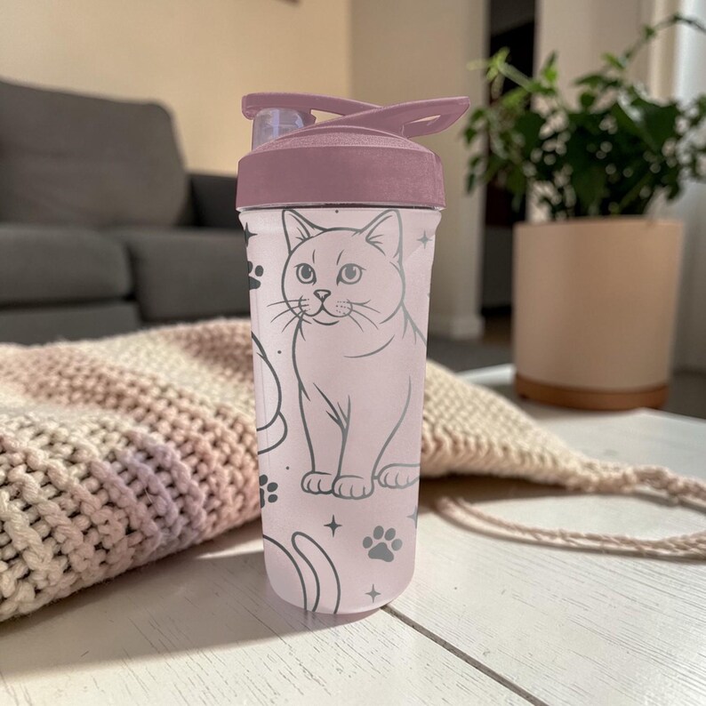 May include: A pink and mauve water bottle featuring a cat illustration. The bottle has a flip-top lid and a light pink body with a cat design, paw prints, and stars. The lid is a darker mauve color.