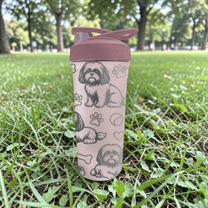 May include: A pink water bottle with a dark pink lid and a flip-top closure. The bottle is decorated with a repeating pattern of Shih Tzu dog illustrations, paw prints, and bones. The bottle is made of plastic and is designed for cold beverages.